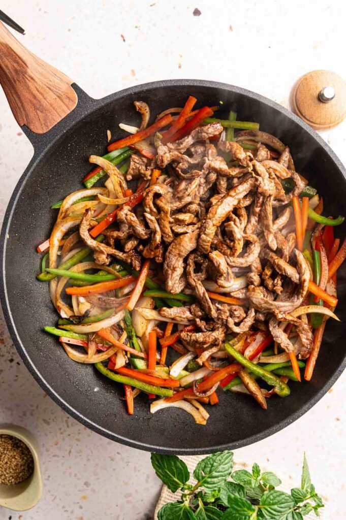 A skillet contains cooked strips of beef, sliced red and green bell peppers, and onions, with steam rising from the mixture&mdash;reminiscent of Panda Express Black Pepper Sirloin Steak.