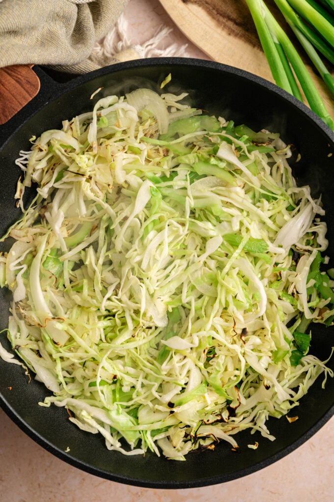 Shredded cabbage saut&eacute;ing in a black skillet with light browning, with fresh green onions on a wooden board beside it.