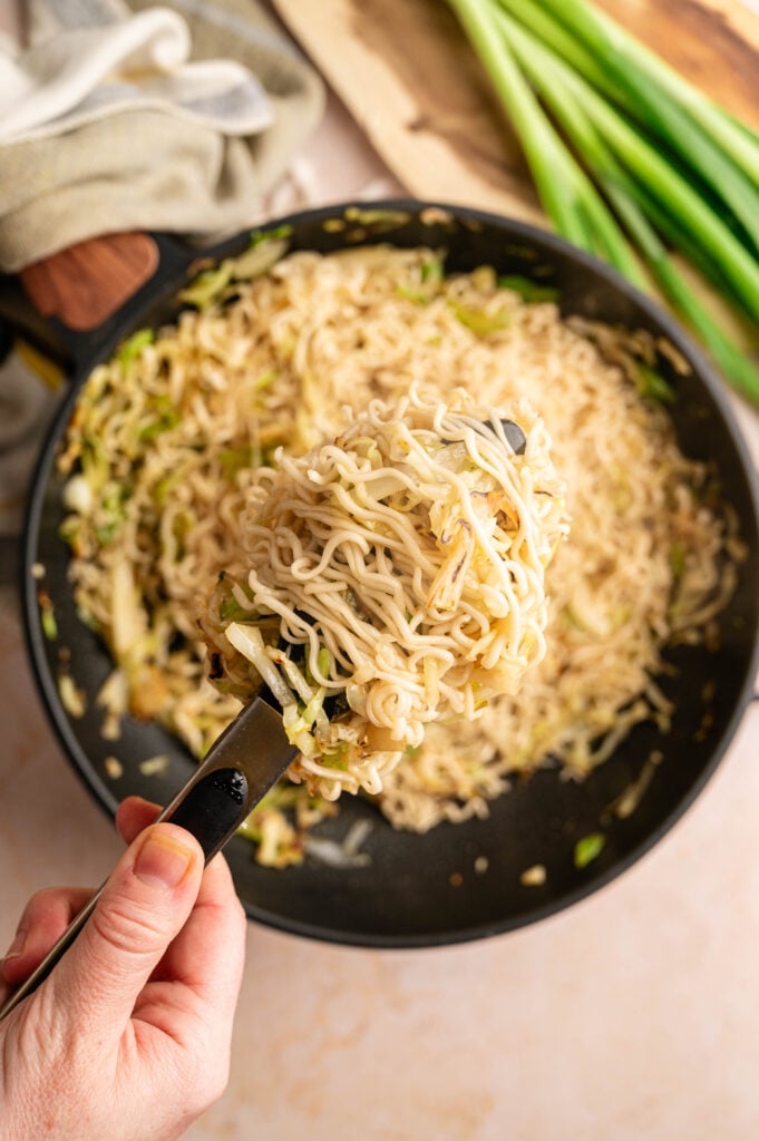 A hand holds up a serving of Panda Express Chow Mein with vegetables over a skillet filled with more noodles; green onions are on the side.