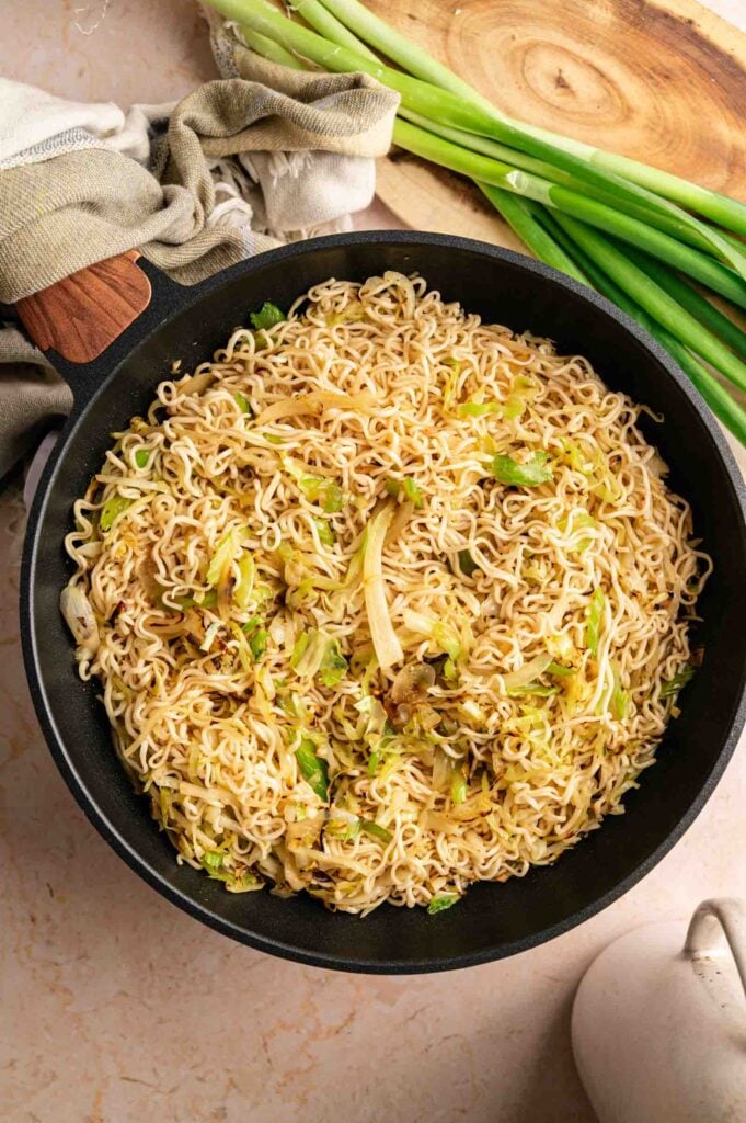 A skillet filled with cooked noodles mixed with chopped cabbage and green onions, reminiscent of Panda Express Chow Mein, is placed on a light countertop with fresh green onions and a cutting board nearby.