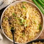 A large bowl of Panda Express Chow Mein with stir-fried cabbage and celery, garnished with sliced green onions and a black fork, served alongside a bunch of whole scallions, a kitchen towel, and a partial view of a second serving in a black pan.