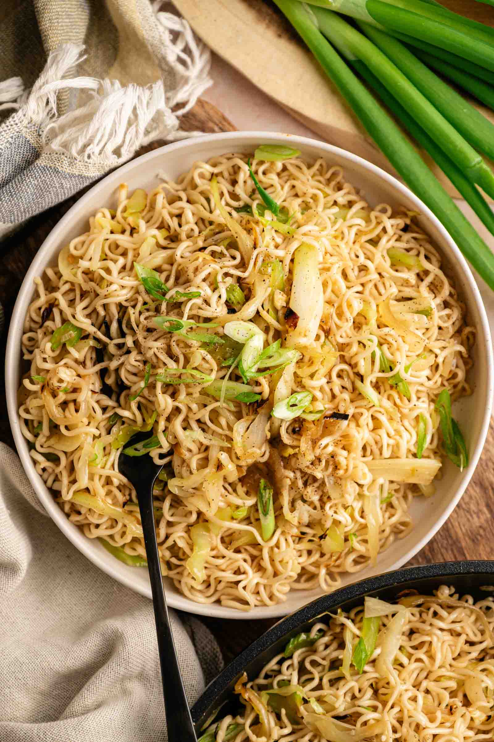 A large bowl of Panda Express Chow Mein with stir-fried cabbage and celery, garnished with sliced green onions and a black fork, served alongside a bunch of whole scallions, a kitchen towel, and a partial view of a second serving in a black pan.