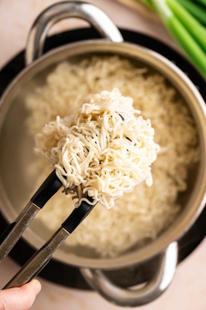 Tongs holding cooked Chow Mein noodles over a pot of boiling water, with uncooked green onions in the background.