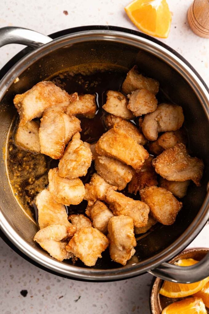 Fried chicken pieces in a pot with savory sauce, reminiscent of Panda Express Orange Chicken. Orange wedges are visible at the top right and bottom right corners of the image.