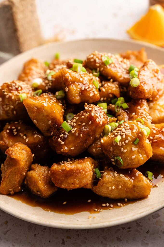 A plate of glazed chicken pieces, inspired by Panda Express Orange Chicken, topped with sesame seeds and chopped green onions, served with a tangy brown sauce.