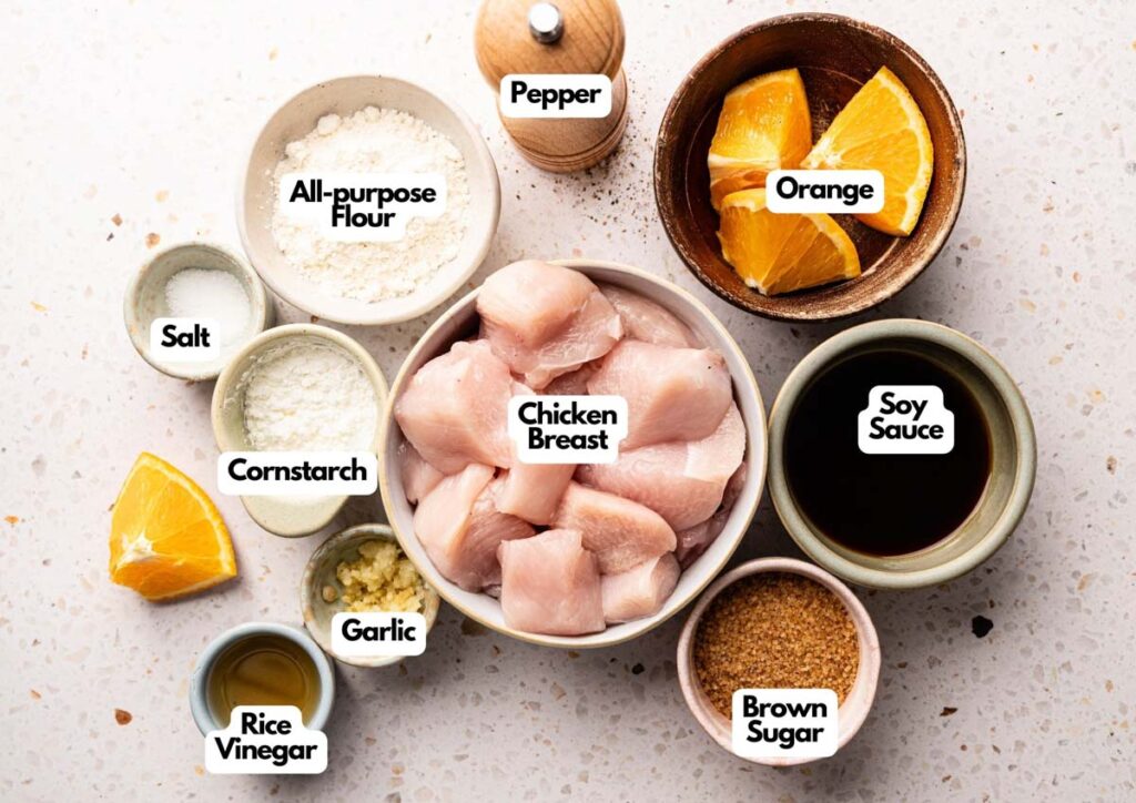 Bowls containing chicken breast, orange slices, soy sauce, brown sugar, garlic, rice vinegar, and other ingredients for Panda Express Orange Chicken are arranged on a speckled surface.
