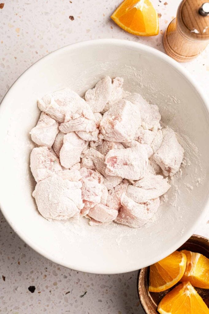 Bite-sized chicken pieces coated in flour and cornstarch in a white bowl, with orange wedges nearby.