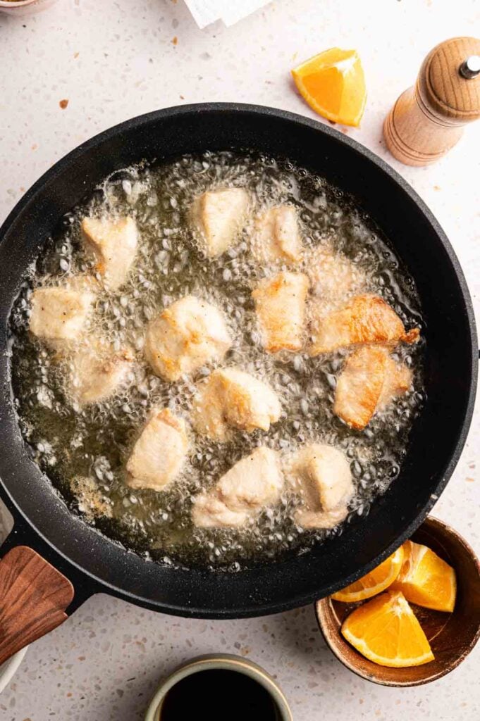 Chunks of battered chicken fry in hot oil in a black skillet, reminiscent of Panda Express Orange Chicken, with orange wedges and a pepper grinder nearby on a light countertop.