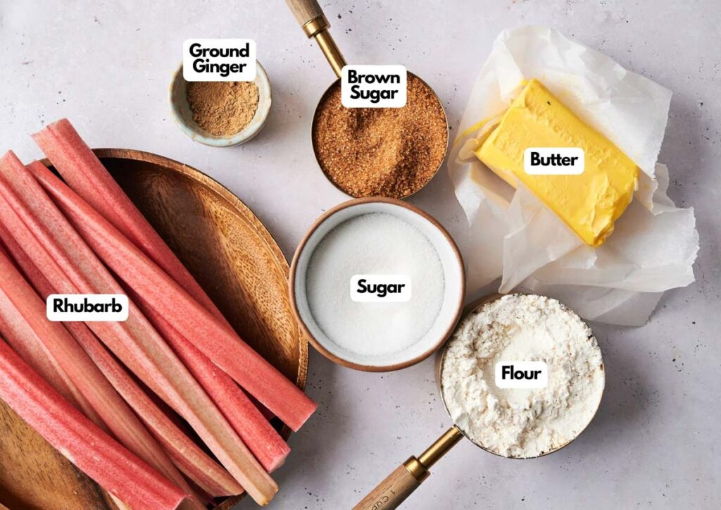 Wooden bowls and measuring cups containing ingredients for Air Fryer Rhubarb Crisp&mdash;rhubarb, ground ginger, brown sugar, sugar, flour, and a block of butter&mdash;are arranged on a light surface, each clearly labeled.