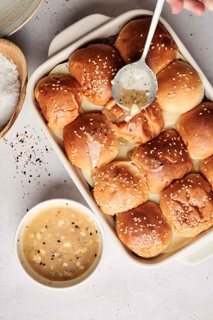 A baking dish filled with sesame-topped bread rolls, some being topped with a ladle of light-colored sauce, ideal for serving Air Fryer Roast Beef Sliders, with a bowl of extra sauce and a bowl of salt beside it.