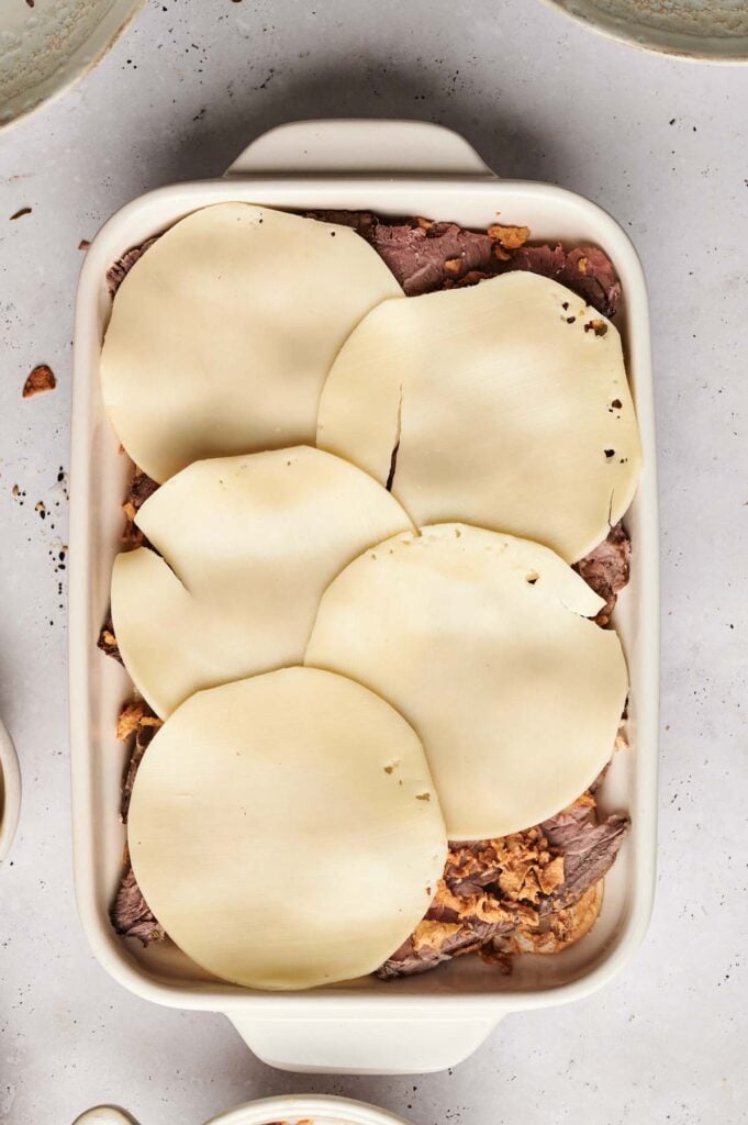 A rectangular baking dish filled with layers of cooked meat, perfect for Air Fryer Roast Beef Sliders, and topped with overlapping slices of provolone cheese.
