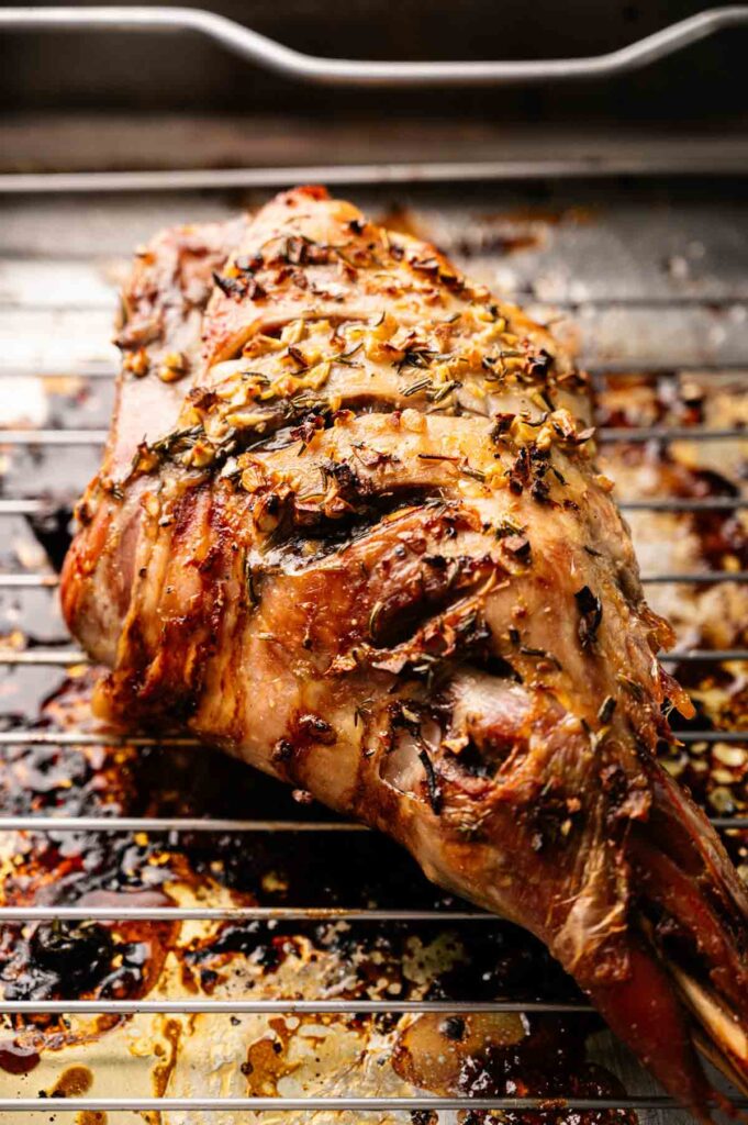 A Slow Roast Leg of Lamb topped with herbs and spices sits on a metal rack in a roasting pan, with browned juices collected below.