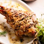 Slow Roast Leg of Lamb garnished with herbs and garlic on a white oval plate, with a side dish of garlic cloves in the background.