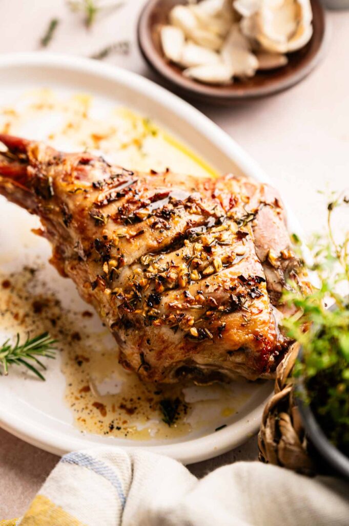 Slow Roast Leg of Lamb garnished with herbs and garlic on a white oval plate, with a side dish of garlic cloves in the background.