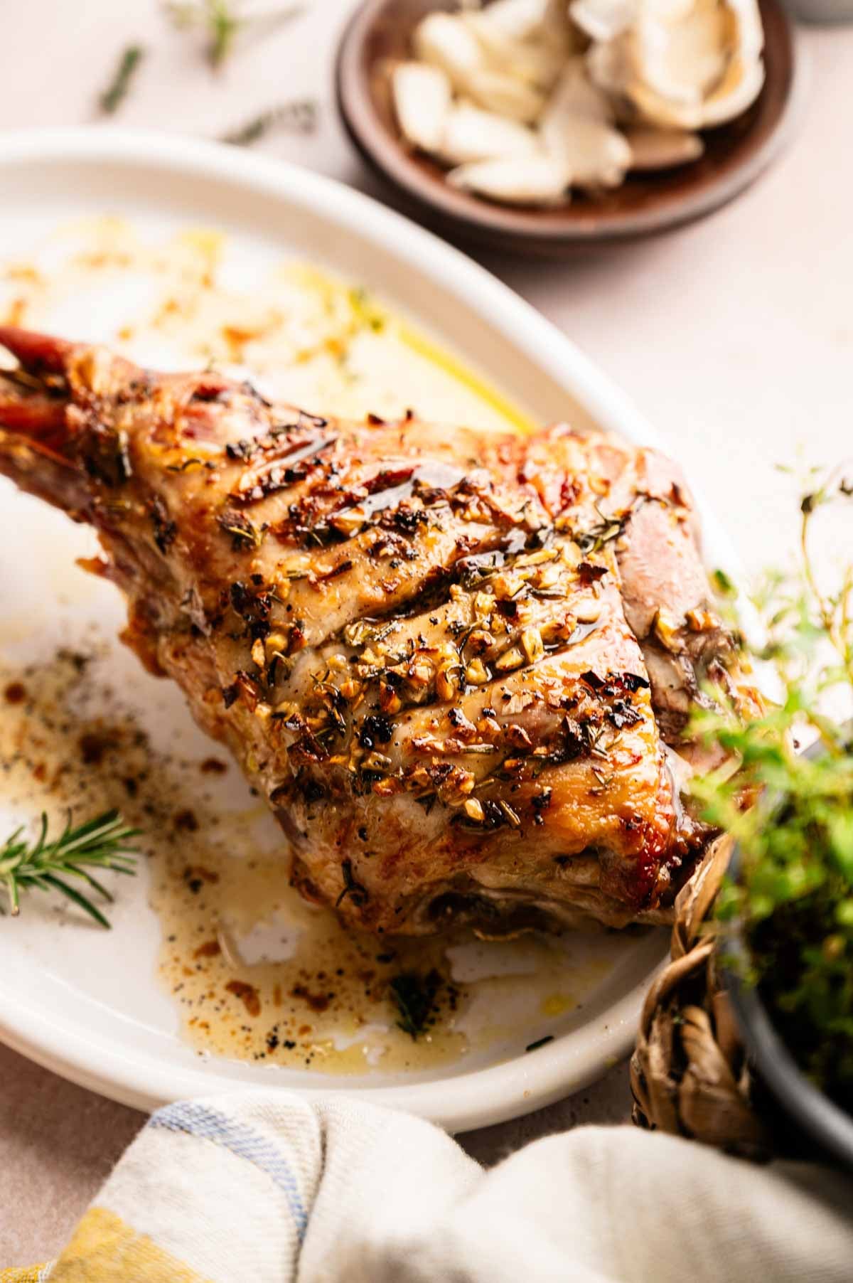 Slow Roast Leg of Lamb seasoned with herbs and garlic on a white platter, accompanied by a side dish of garlic cloves and fresh rosemary nearby.