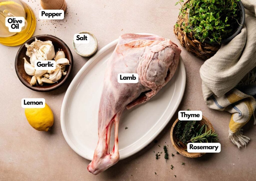 A raw Slow Roast Leg of Lamb sits on a white platter, surrounded by bowls of olive oil, salt, pepper, garlic, lemon, fresh thyme, and rosemary.