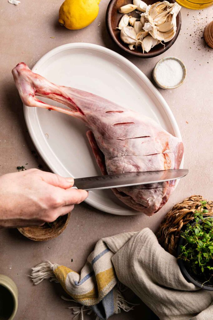 A hand slicing small slits into a raw leg of lamb on a white platter.