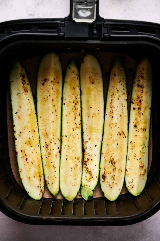 Six seasoned zucchini halves are arranged in a single layer inside an air fryer basket, perfect for making delicious Air Fryer Baked Zucchini.