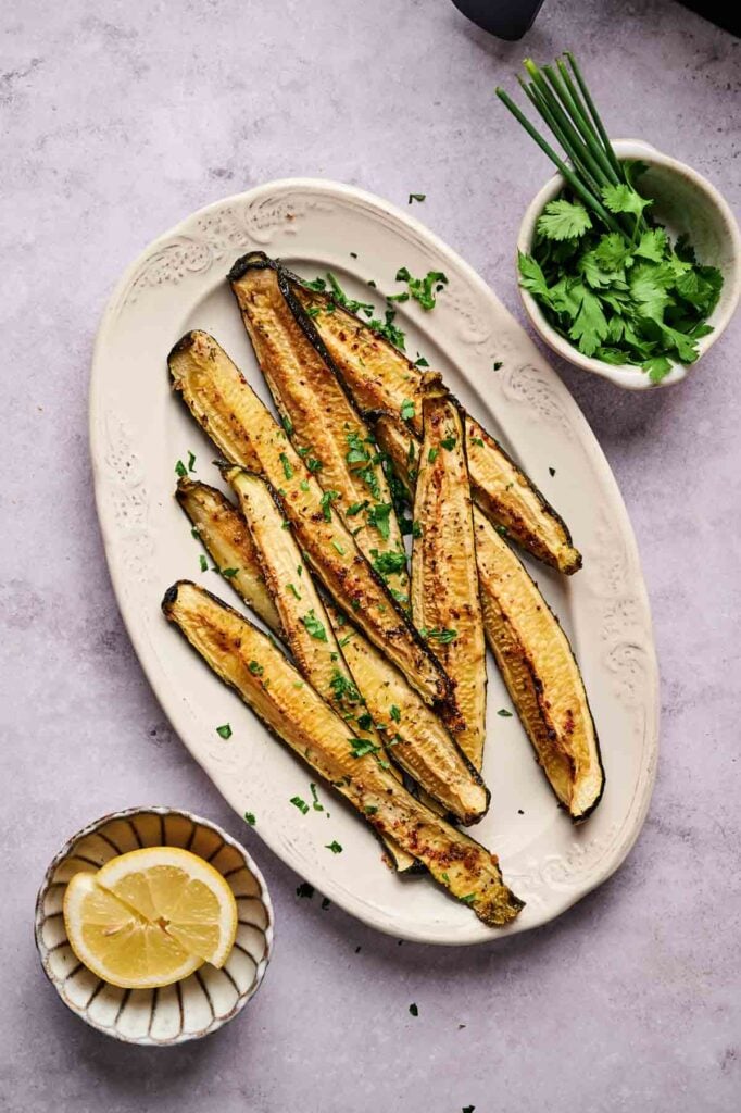 Air Fryer Baked Zucchini spears garnished with chopped herbs on an oval plate, with bowls of fresh cilantro, chives, and lemon wedges nearby.