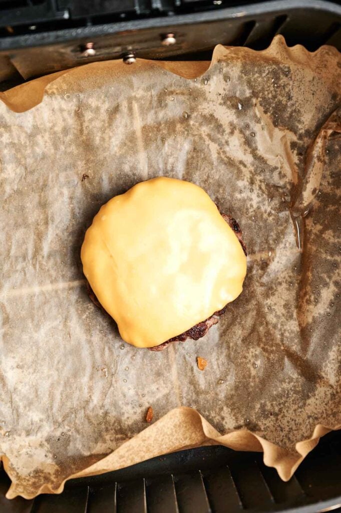 An Air Fryer Cheeseburger patty with melted cheese sits on brown parchment paper in a baking tray.