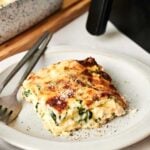 A slice of baked Air fryer Chicken Alfredo Lasagna is served on a white plate with a fork and knife, with a baking dish and air fryer in the background.