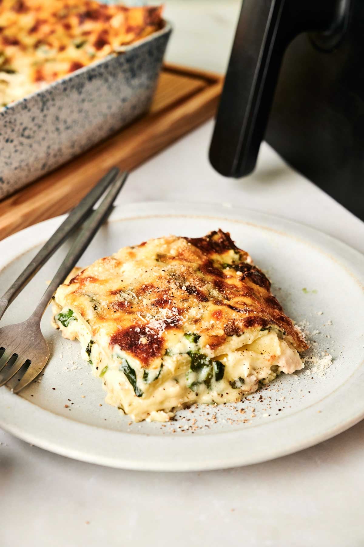 A slice of baked Air fryer Chicken Alfredo Lasagna is served on a white plate with a fork and knife, with a baking dish and air fryer in the background.