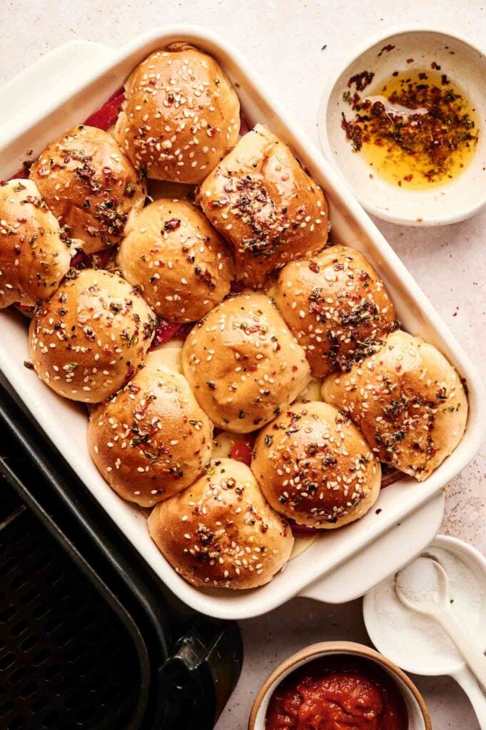 A baking dish filled with golden Air Fryer Italian Sliders topped with sesame seeds and fresh herbs, next to bowls of oil, sauce, and a sleek black air fryer.