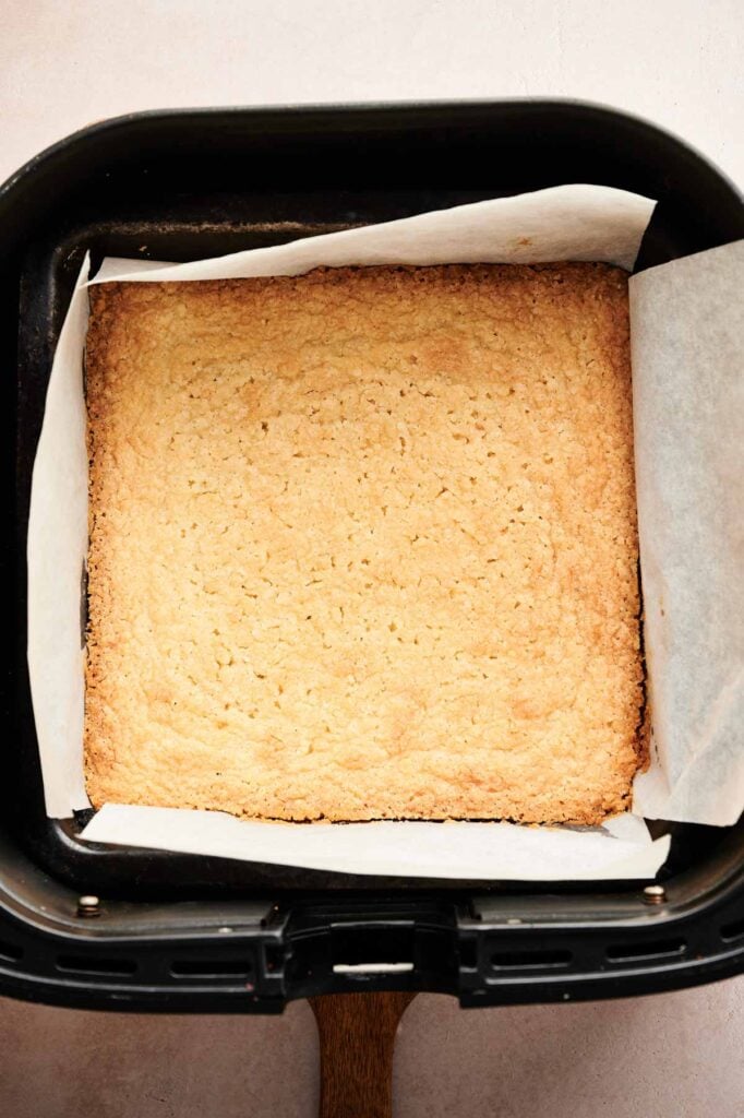 A square, lightly golden-baked Air Fryer Pecan Pie Bar crust sits in a parchment-lined air fryer basket.