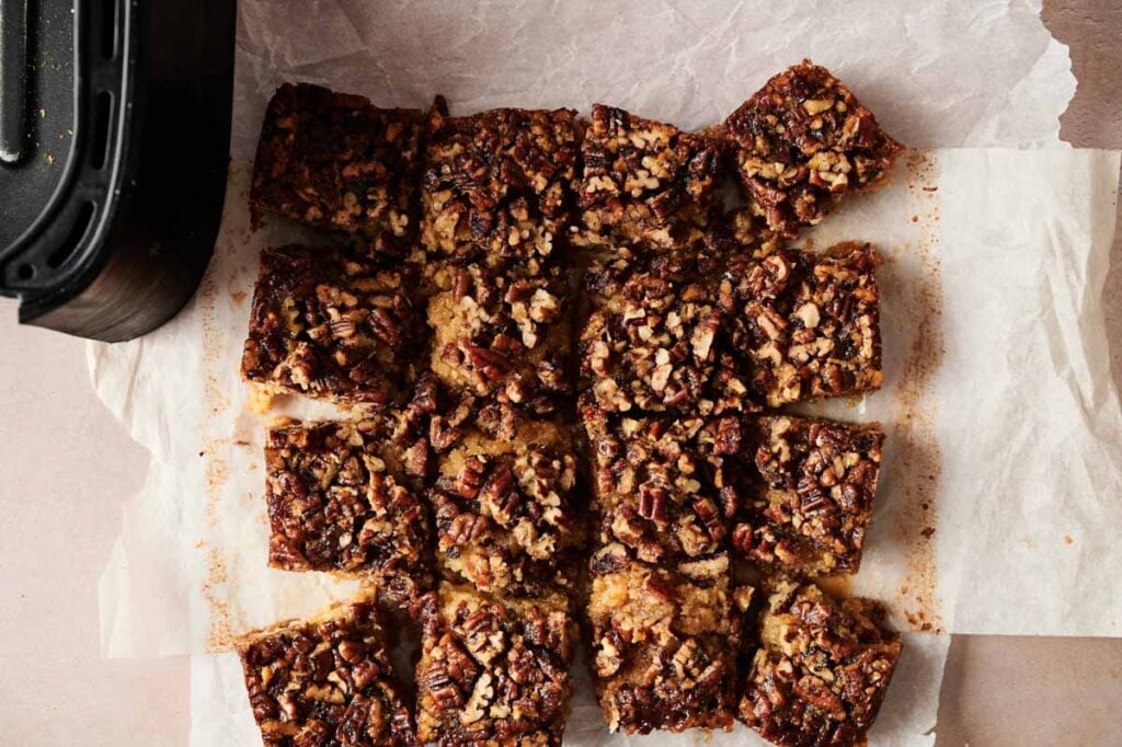 A batch of Air Fryer Pecan Pie Bars cut into squares on parchment paper, with part of a baking pan visible in the corner.