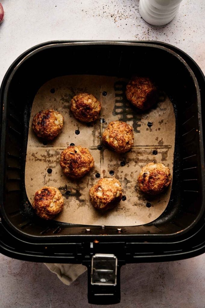 Seven cooked Air Fryer Turkey Meatballs rest on parchment paper inside an air fryer basket, with a few visible grease spots on the paper.