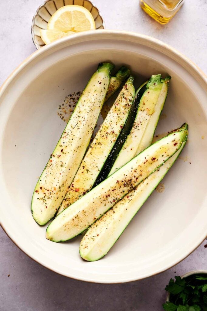 Four halved zucchinis seasoned with spices in a large white bowl, perfect for making Air Fryer Baked Zucchini, with a dish of lemon slices and a bottle of oil nearby.