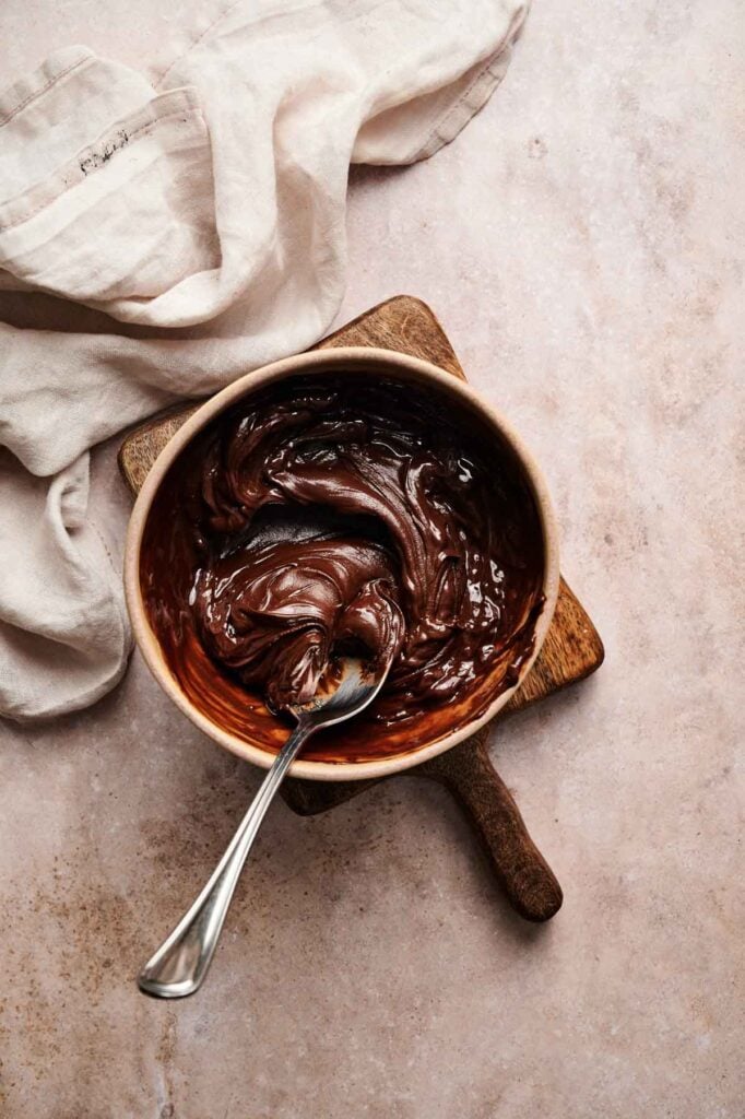 A bowl of melted chocolate with a spoon resting inside, placed on a wooden board next to a beige cloth on a marble surface.