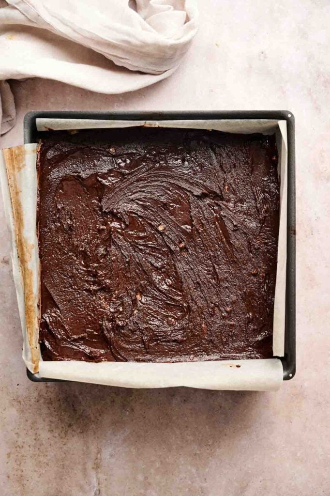 A square pan lined with parchment paper filled with Buckeye Brownies, topped with a smooth layer of melted chocolate spread evenly over the surface.