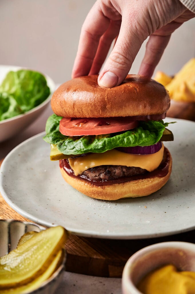 A hand holding an Air Fryer Cheeseburger with lettuce, tomato, pickles, onion, and cheese on a plate, with bowls of pickles and lettuce in the background.