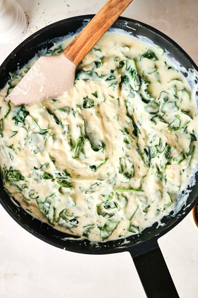 A black skillet filled with creamy spinach sauce, perfect for layering in Air Fryer Chicken Alfredo Lasagna, being stirred with a beige spatula on a white surface.