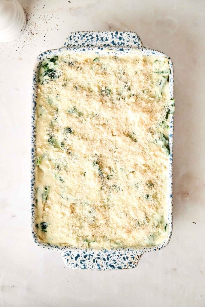 A rectangular baking dish filled with an unbaked creamy Air Fryer Chicken Alfredo Lasagna, topped with grated cheese and breadcrumbs, with green vegetable pieces visible.