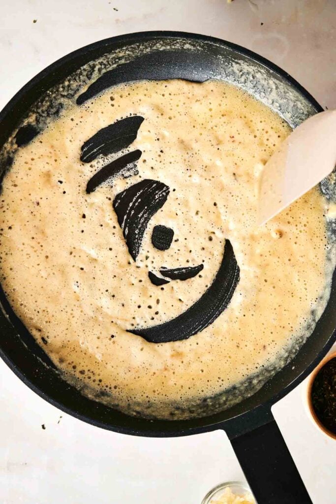 A spatula stirs a bubbly, light brown sauce in a black skillet on a white surface, perfect for making creamy Air Fryer Chicken Alfredo Lasagna.