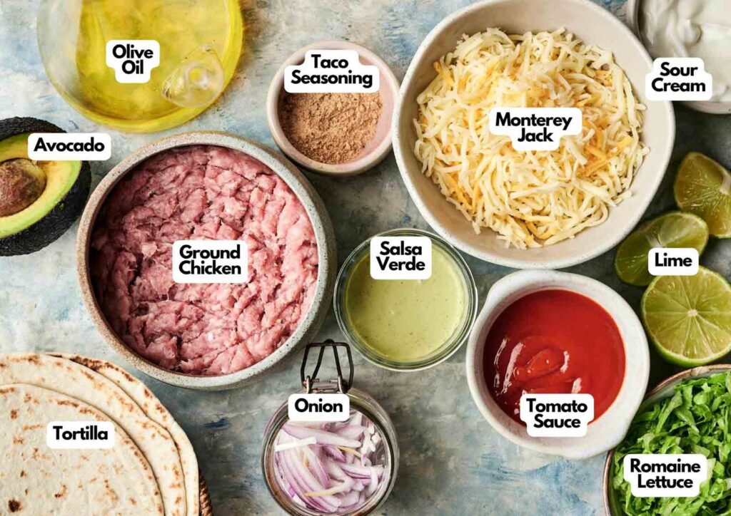Top-down view of various Air Fryer Chicken Taco ingredients in bowls and plates, including ground chicken, tortilla, avocado, olive oil, taco seasoning, salsa verde, onion, Monterey Jack cheese, tomato sauce, sour cream, lime, and romaine lettuce.