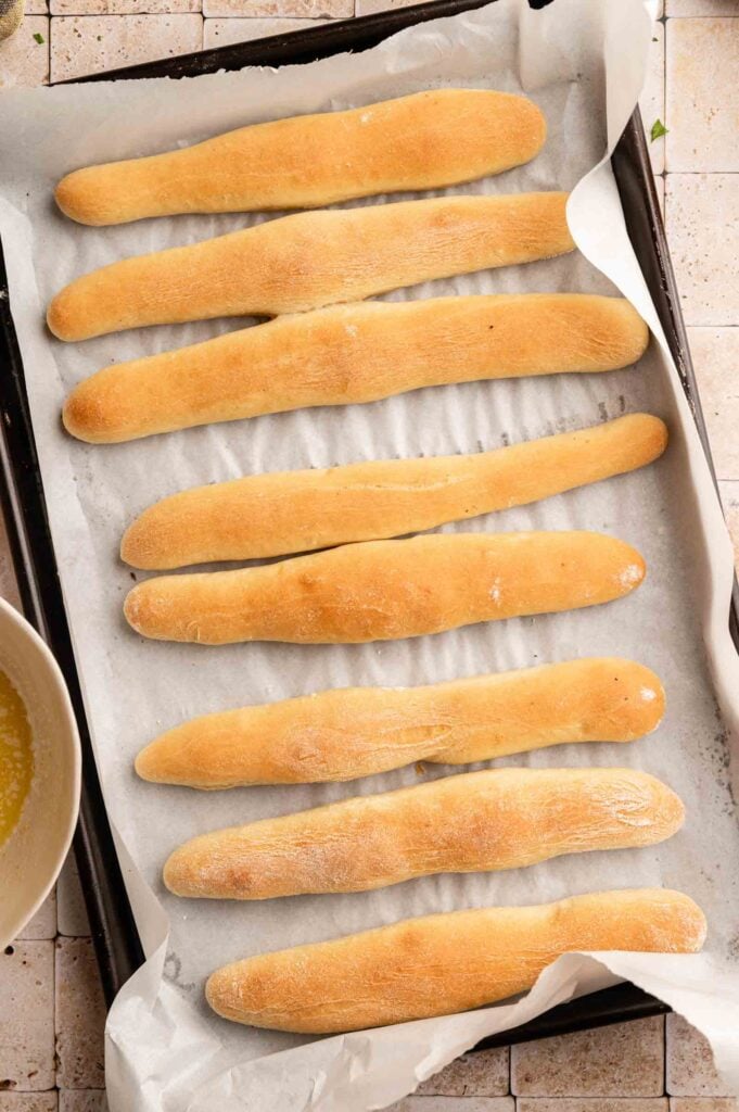 Eight Copycat Olive Garden Breadsticks are arranged in rows on a parchment-lined baking sheet.