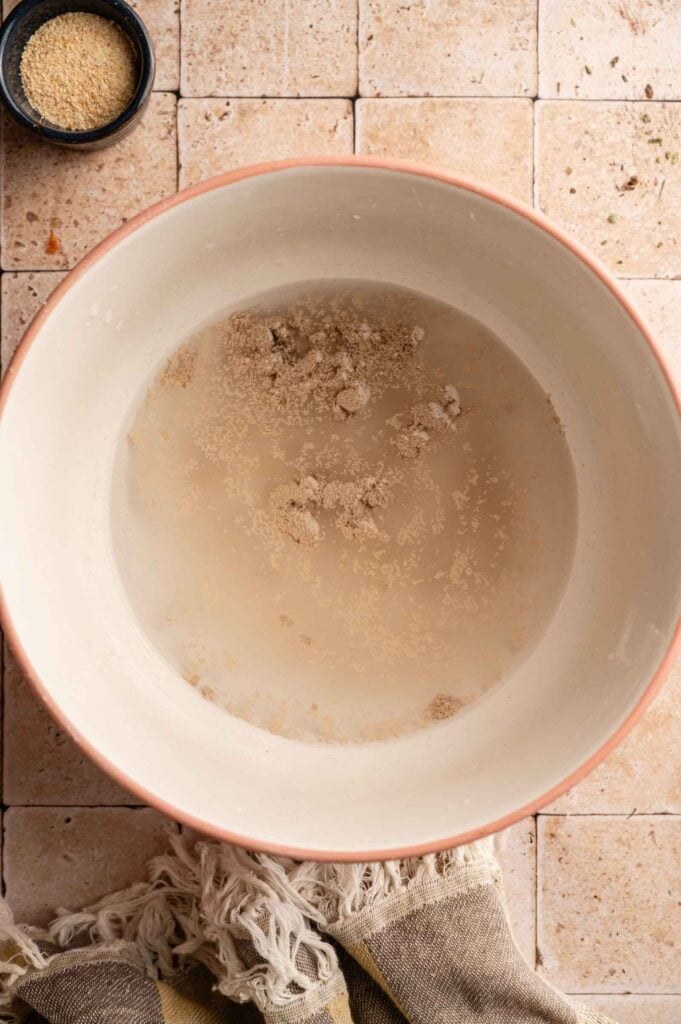 A large bowl with water and dry yeast sprinkled on top sits on a tiled surface, ready for making Copycat Olive Garden Breadsticks, with a small bowl of granulated ingredient nearby and a cloth partially visible.