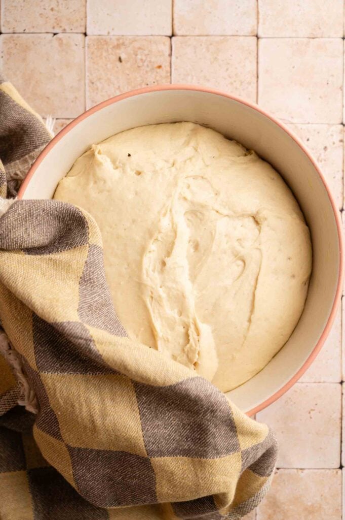 A bowl of risen Copycat Olive Garden Breadsticks dough is partially covered with a brown and yellow checkered cloth on a tiled surface.