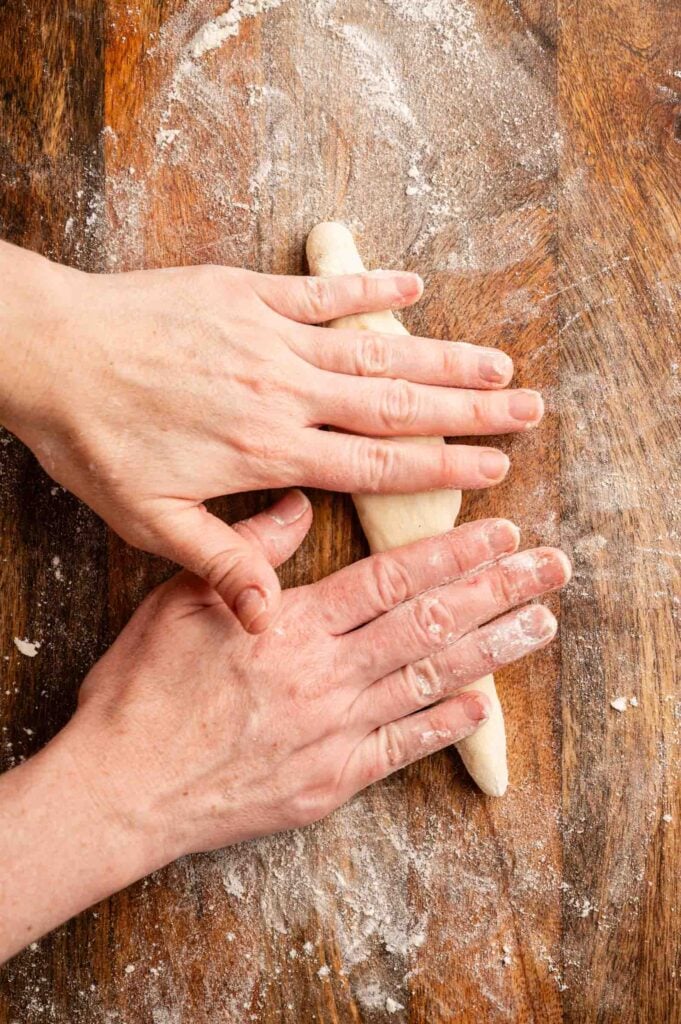 Hands rolling dough into a long shape on a floured wooden surface, preparing homemade copycat Olive Garden breadsticks.