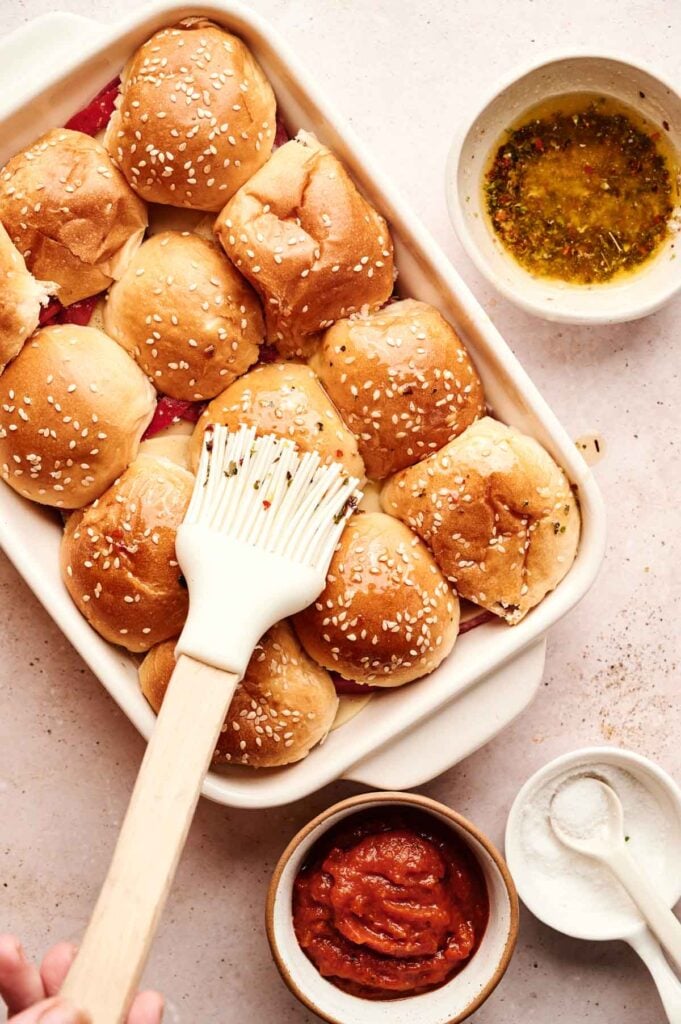 A baking dish of Air Fryer Italian Sliders being brushed with melted butter, surrounded by bowls of sauce, seasoning, and melted butter on a light surface.