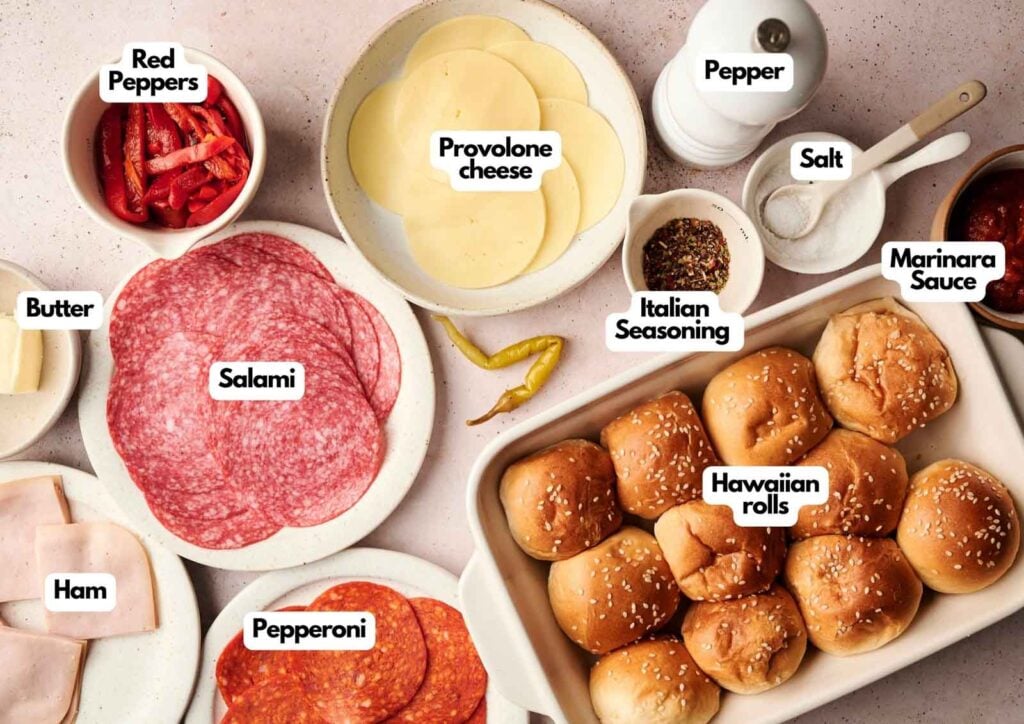 Ingredients for making Air Fryer Italian Sliders are arranged on a countertop, including Hawaiian rolls, salami, pepperoni, ham, provolone cheese, red peppers, butter, Italian seasoning, salt, pepper, and marinara sauce.