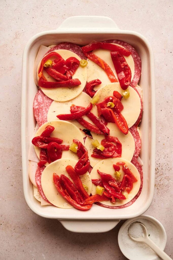 A white baking dish filled with layered slices of salami, provolone cheese, and strips of red peppers, perfect for making Air Fryer Italian Sliders, with a small dish of salt beside it.