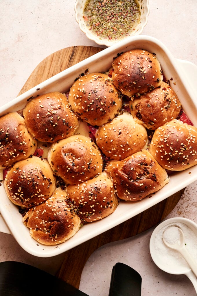 A baking dish filled with twelve golden brown dinner rolls, perfect for serving Air Fryer Italian Sliders, topped with sesame and poppy seeds, with bowls of seasoning and salt nearby.