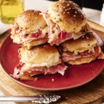 A plate of four stacked Air Fryer Italian Sliders filled with layers of deli meat and melted cheese, set on a red plate with a bottle of olive oil in the background.