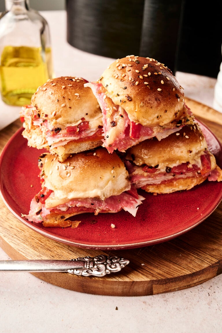 A plate of four stacked Air Fryer Italian Sliders filled with layers of deli meat and melted cheese, set on a red plate with a bottle of olive oil in the background.