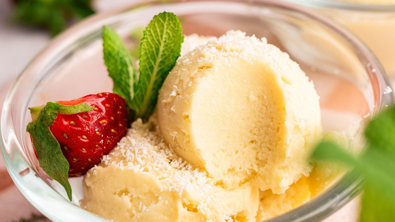 A glass bowl with two scoops of vanilla ice cream, garnished with shredded coconut, a fresh strawberry, and a sprig of mint.
