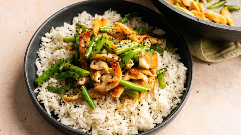 A bowl of white rice topped with stir-fried chicken, green beans, and black sesame seeds.
